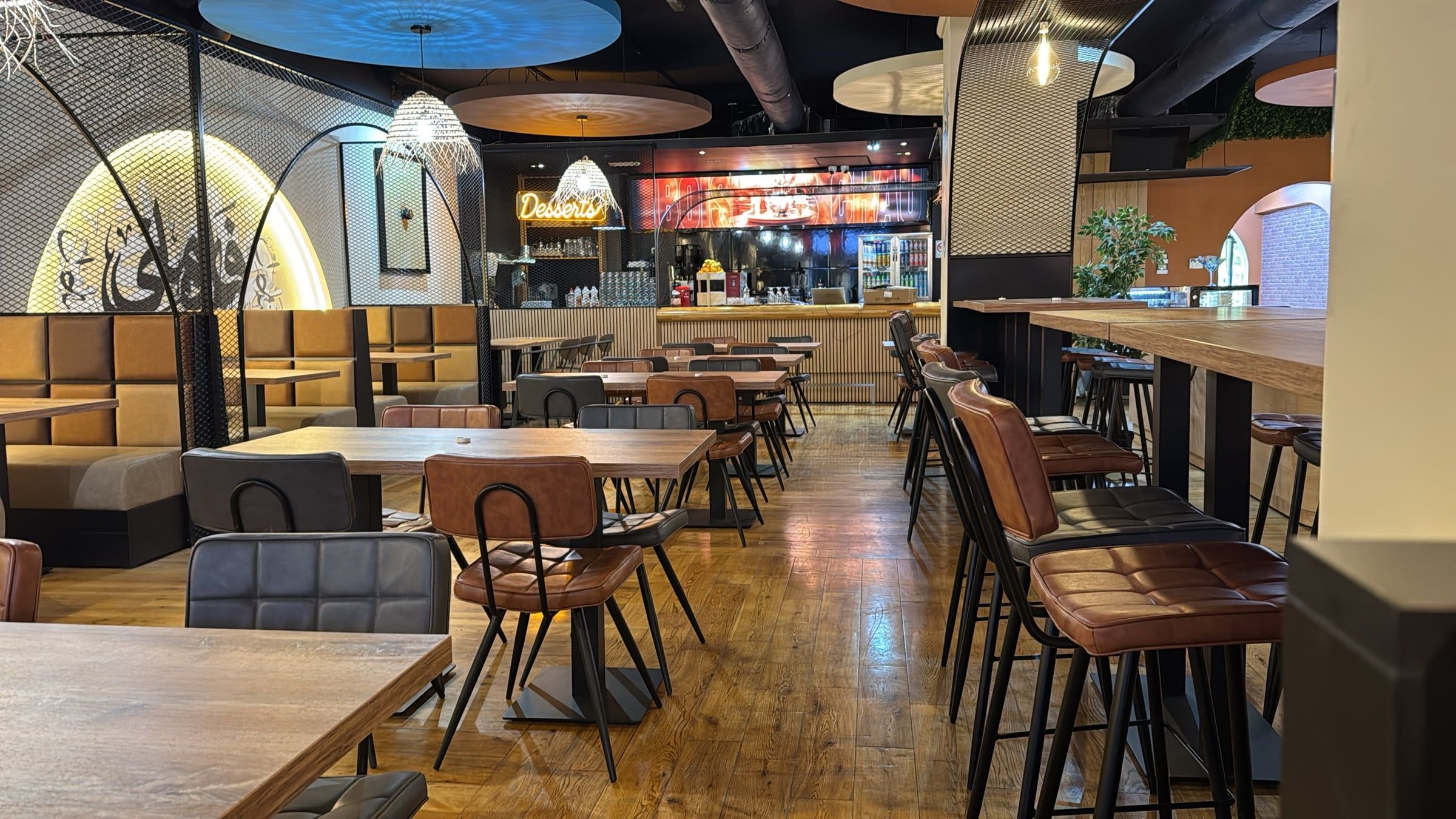 La grande salle du FAMI Café : tables en bois, chaises en cuir, suspensions tressées et comptoir des desserts au fond